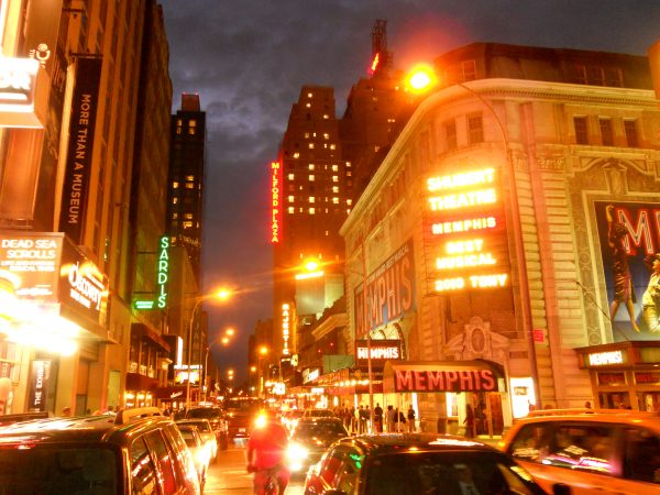 Cars and pedestrians move along a lively Broadway street at dusk, with bright neon signs and Shubert Theatre showing MEMPHIS.