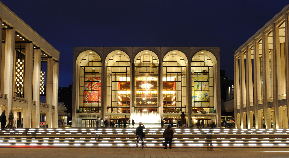 Lincoln Center glows at night in New York City, featuring arched windows, colorful banners, a lit fountain, and gathering visitors.