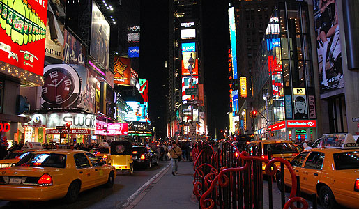 Crowds fill Times Square at night in New York City, surrounded by colorful billboards, glowing Broadway theaters, and yellow taxis.