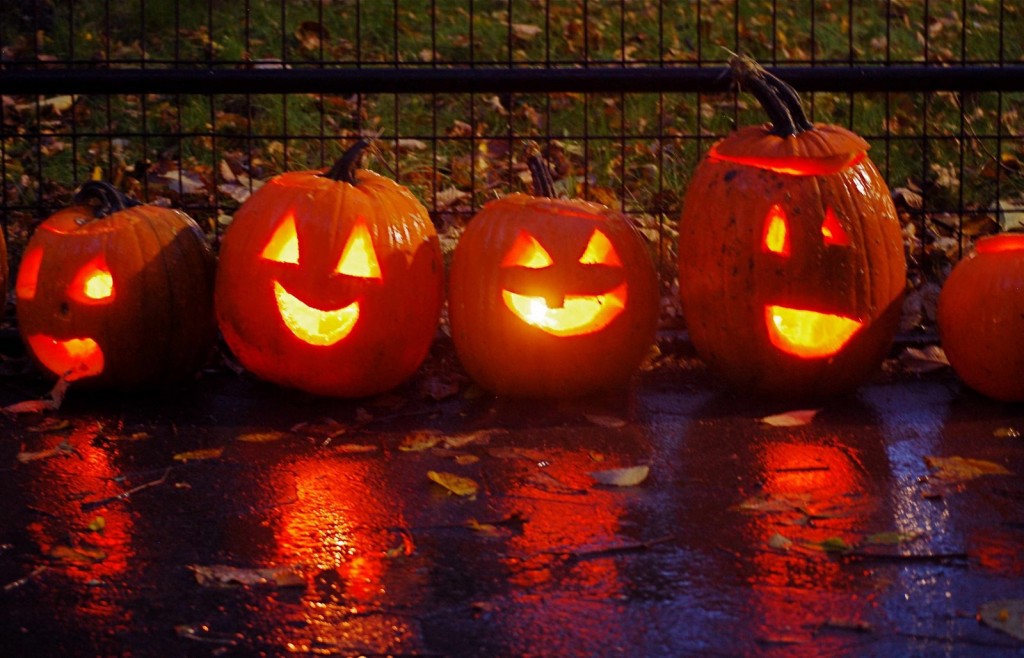 Four carved jack-o’-lanterns with unique faces rest on wet pavement near a fence, surrounded by autumn leaves in New York City.
