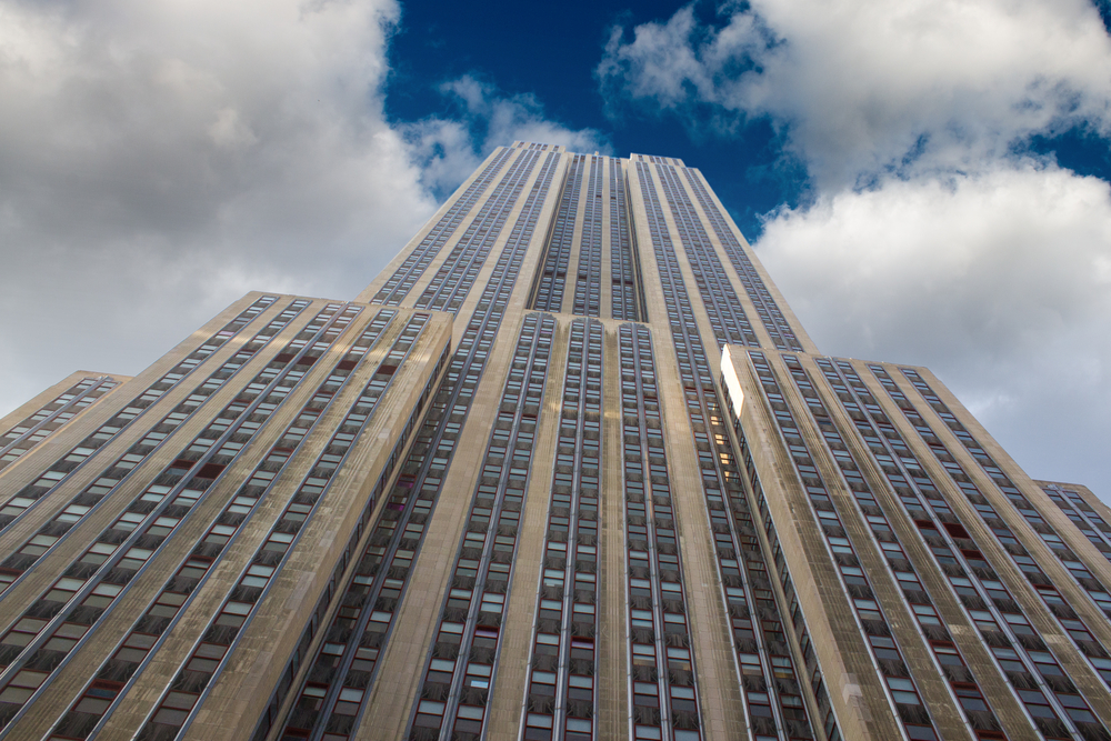 Looking up from the base of the Empire State Building in New York City, its many windows and stone facade reach into blue sky.