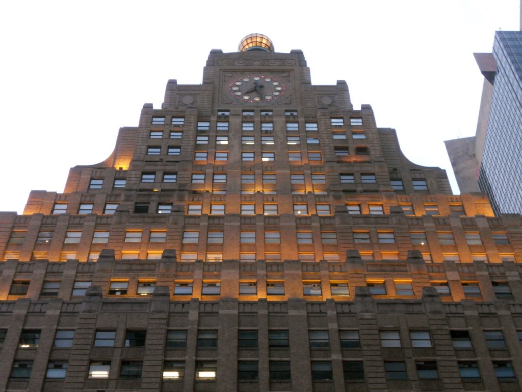 A stepped Art Deco skyscraper with rows of glowing windows and a clock near its top towers over Times Square, New York.