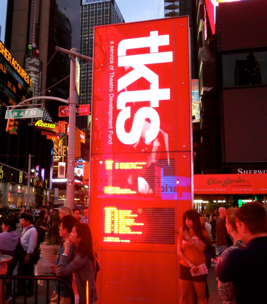 A bright red TKTS booth sign in Times Square, New York City, attracts crowds strolling among glowing billboards at dusk.