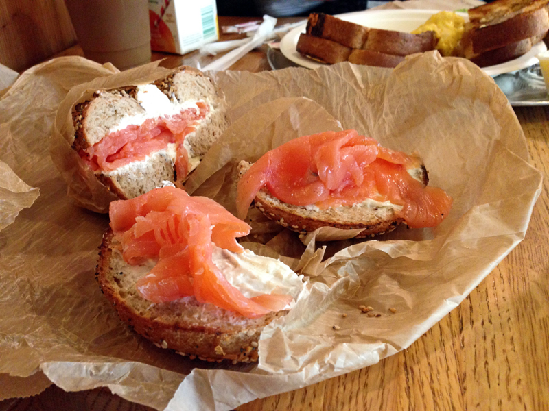 A toasted bagel topped with cream cheese and smoked salmon rests on brown parchment atop a wooden table in New York.