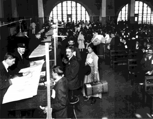 A group of immigrants wait in a hall with arched windows on Ellis Island as officials check their papers and belongings.