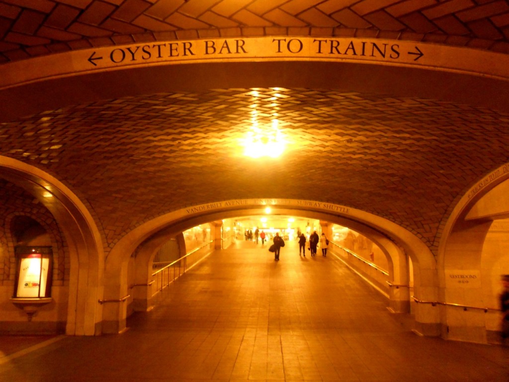 A warmly lit arched hallway in Grand Central Terminal, New York, guides people beneath a sign reading Oyster Bar to Trains. Grand Central Whispering Gallery.