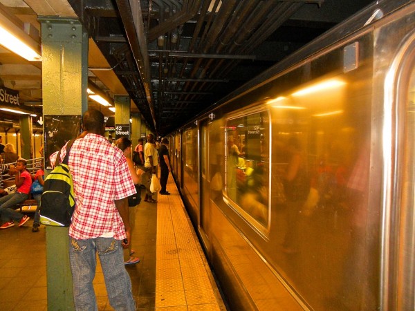 A man in a red and white plaid shirt waits on the 42nd Street subway platform as a train approaches amid bustling commuters.