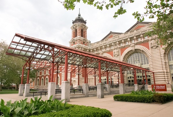 The historic Ellis Island Immigration Museum features red brick, arched windows, a clock tower, nearby trees, and the Ellis Island ferry landing.