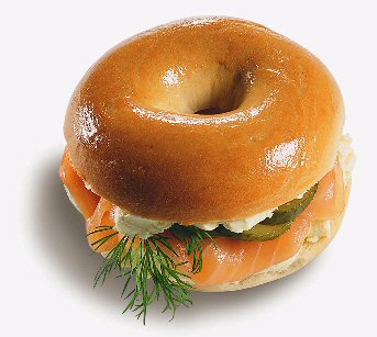 A classic New York bagel sandwich stacked with smoked salmon, cream cheese, cucumber slices, and fresh dill sits on a white surface.