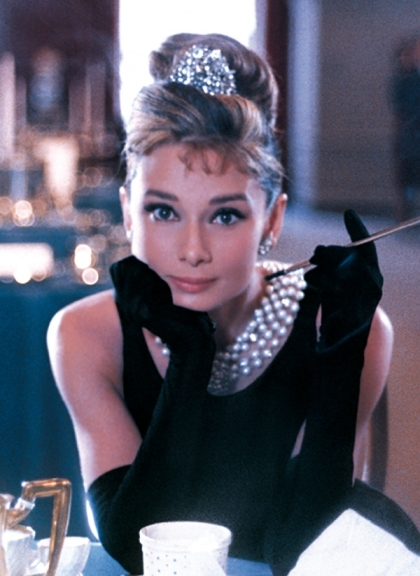 Audrey Hepburn, dressed as Holly Golightly, sits at a table in Tiffany’s, holding a cigarette holder and gazing forward.
