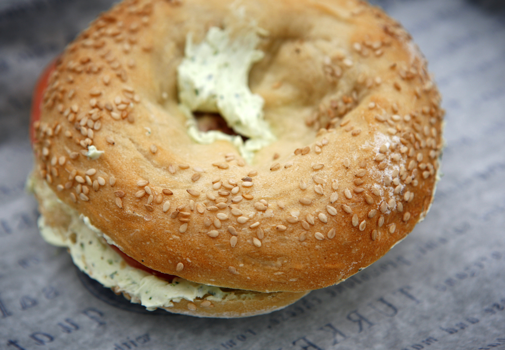 A New York sesame bagel stuffed with herbed cream cheese and tomato sits on parchment paper, ready to eat.