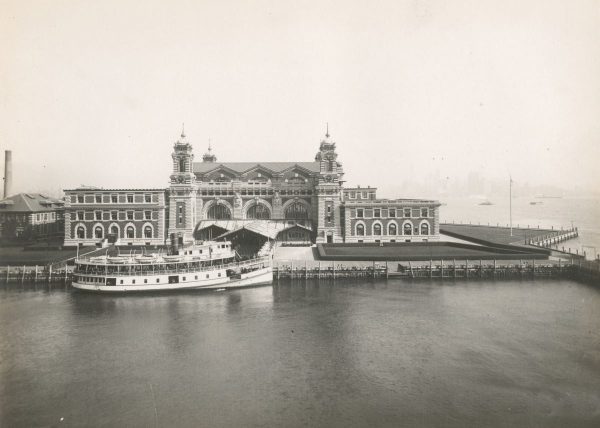 Ellis Island’s main building stands by the harbor in New York, with a ferry docked and Manhattan’s skyline faintly visible.