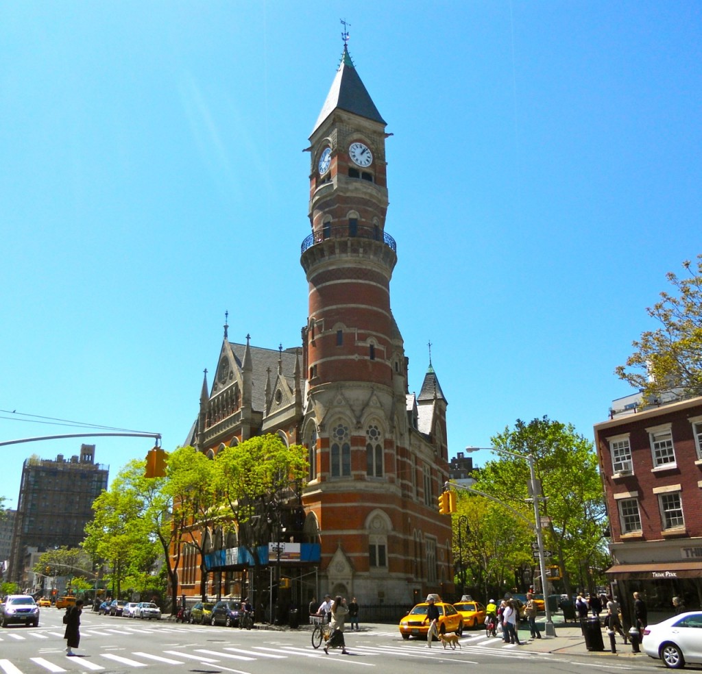 A red-brick Jefferson Market Clock Tower rises above a busy Greenwich Village intersection, surrounded by pedestrians, cyclists, yellow taxis, and trees.