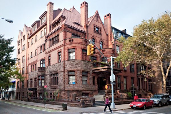 Pedestrians walk past a historic Brooklyn Heights brownstone, surrounded by trees, parked cars, and streetlights on a sunny day.