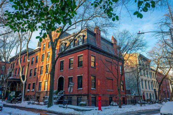 A red brick townhouse in Brooklyn Heights stands on a snowy corner, sunlight casting long shadows among historic buildings and bare trees.