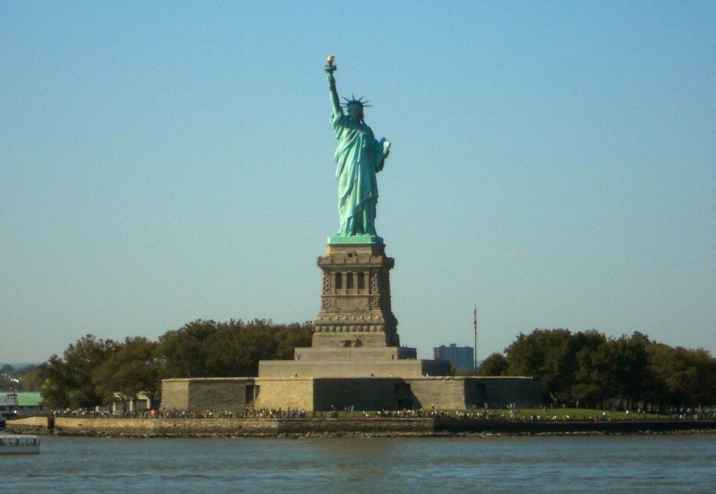 The Statue of Liberty rises above Liberty Island in New York Harbor, encircled by water and greenery under a bright blue sky.