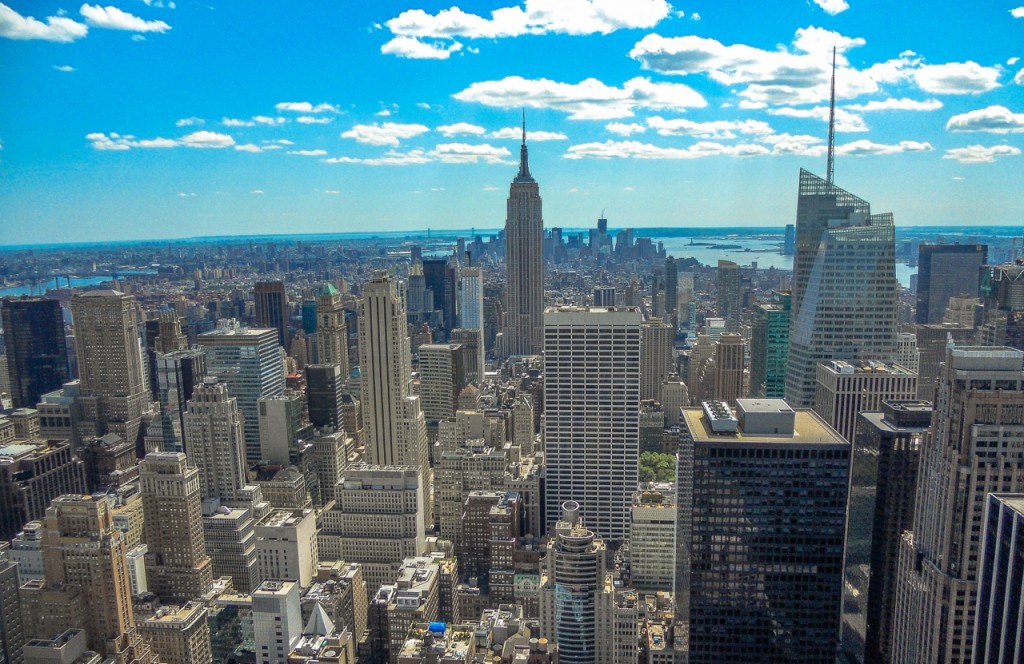 The Empire State Building rises among Manhattan skyscrapers on a clear day, with blue sky and scattered clouds overhead.