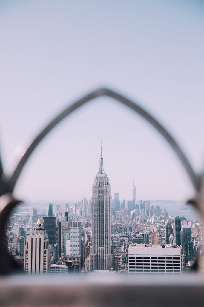 The Empire State Building rises above New York City’s skyline, framed by a blurred archway and set against a clear sky.
