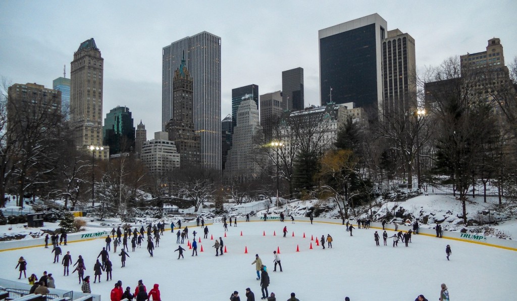 People ice skate in Central Park, New York, surrounded by snow-covered trees and skyscrapers beneath a cloudy winter sky.