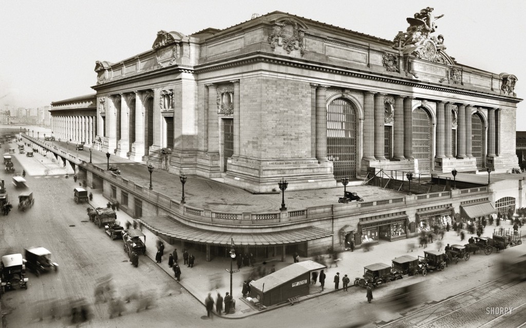 Grand Central Station in New York City bustles with early 1900s cars, horse-drawn carriages, and pedestrians near its grand entrance.