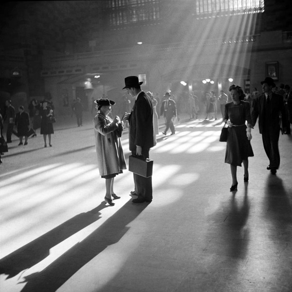 Several people in vintage clothing walk through Grand Central Terminal, bathed in sunlight streaming through tall windows, creating dramatic shadows.