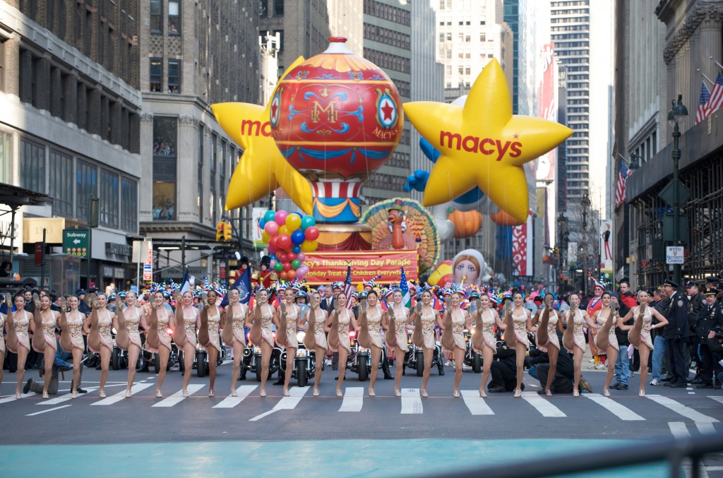 Several dancers in matching costumes perform energetically on a New York City street during the Macy’s Thanksgiving Day Parade, with giant balloons behind.