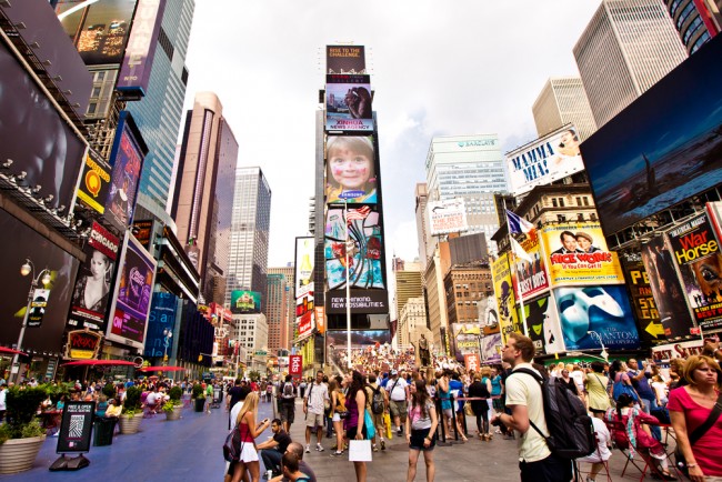 A diverse crowd gathers in Times Square, New York City, surrounded by tall buildings and bright electronic billboards under scattered clouds.