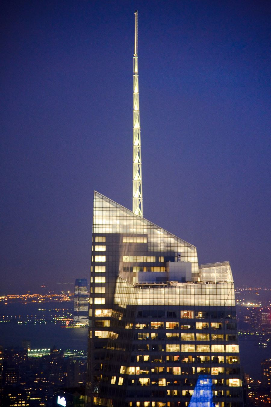 A brightly lit modern skyscraper with a tall spire stands in New York City, surrounded by glowing lights and distant buildings.