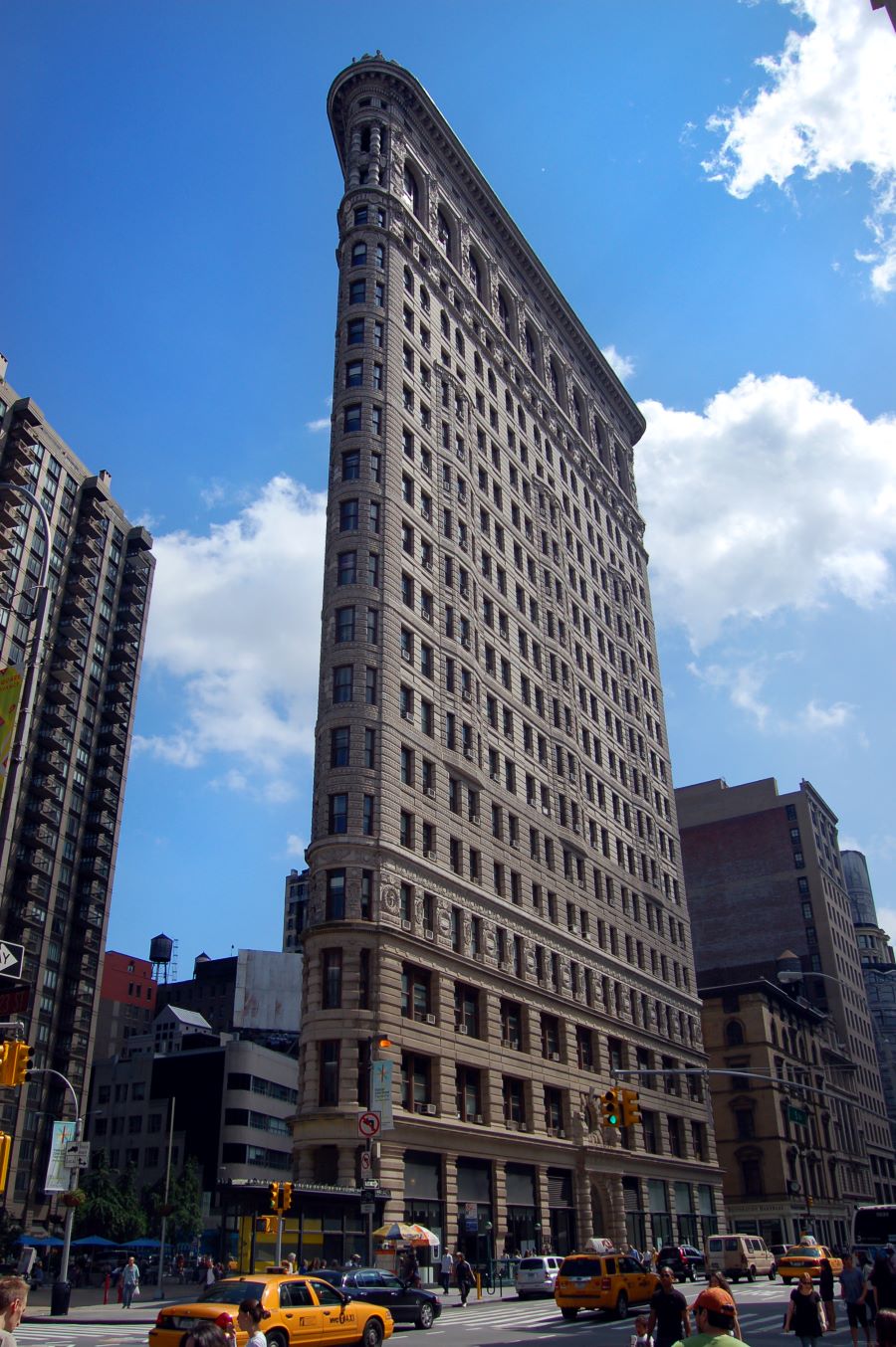 The Flatiron Building rises on a bustling New York City corner, framed by yellow taxis and neighboring skyscrapers beneath a partly cloudy sky.