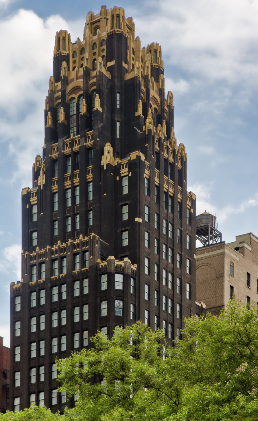 A tall Art Deco skyscraper with ornate gold accents rises above green trees in New York City, under a cloudy sky.