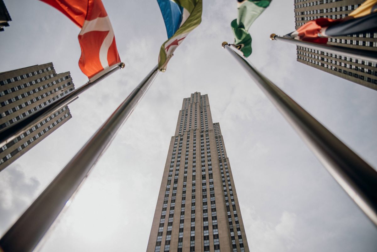 Several international flags wave on flagpoles, with tall New York City skyscrapers towering upward under a cloudy sky in Rockefeller Center.