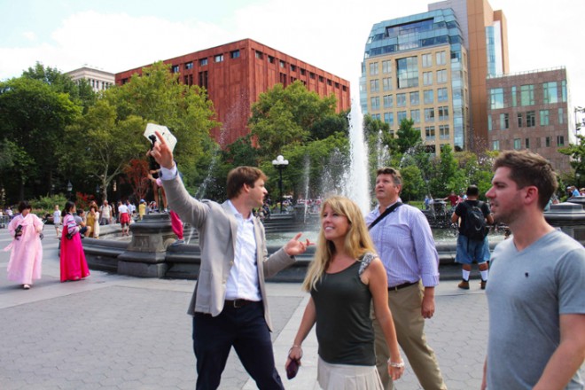 A man takes a selfie with friends by a large city fountain in Central Park, surrounded by tall buildings and green trees.