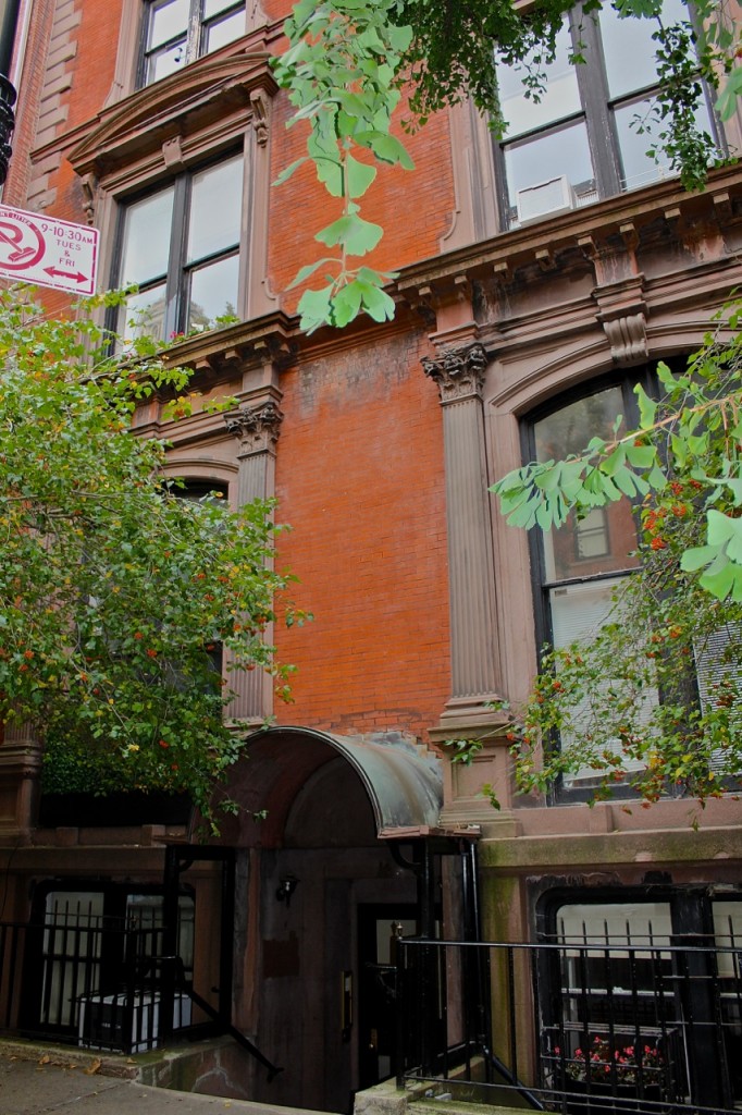 A red brick building with stone columns stands behind leafy trees in New York, its entrance partially hidden and marked by a No Parking sign.