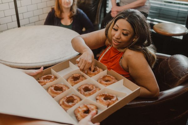 A woman on a brown leather chair reaches for a large glazed donut during a Lower East Side food tour with friends.