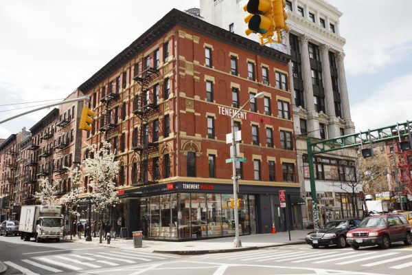 A red-brick tenement building with black fire escapes and a TENEMENT sign stands on a bustling Lower East Side intersection.