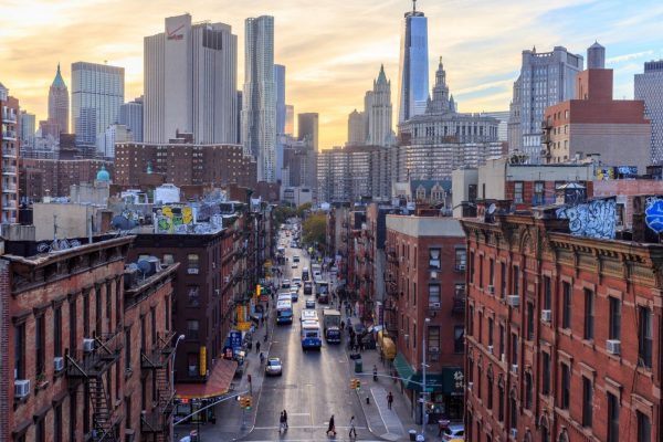 Brick buildings line a busy Lower East Side street in New York as people walk and vehicles travel toward glowing skyscrapers at sunset.