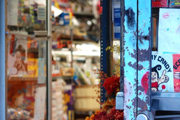 A weathered blue door opens into a bright Lower East Side convenience store filled with colorful products, autumn leaves, and vintage signage.