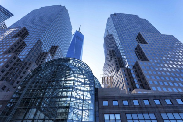 Tall modern skyscrapers with reflective glass, a curved glass atrium in front, and clear blue sky suggest One World Trade Center.