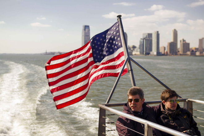 Two people sit on a boat deck near an American flag, cruising toward the Statue of Liberty with New York City behind them.