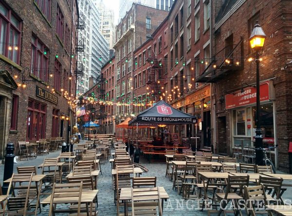 Empty wooden tables and chairs sit on a cobblestone street in Lower Manhattan, with string lights glowing above restaurant patios.