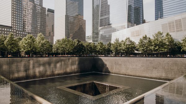 Water pours into a square void at the 9/11 Memorial in Lower Manhattan, bordered by trees and tall modern buildings.