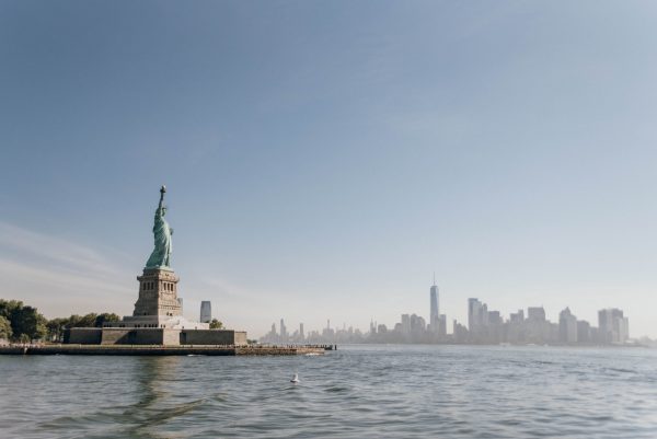 The Statue of Liberty rises above Liberty Island, with surrounding water and the hazy New York City skyline in the background.