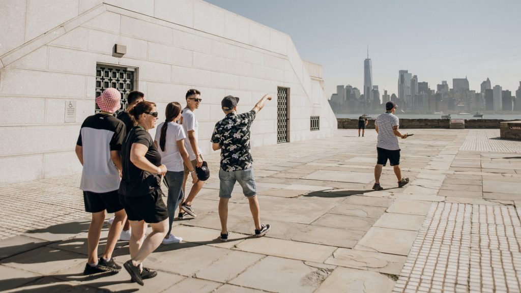 Several people walk on a stone terrace as one points at New York City’s skyline; another stands alone gazing toward Manhattan.