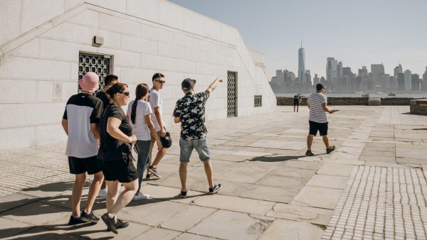 Several people walk on a stone terrace as one points at New York City’s skyline; another stands alone gazing toward Manhattan.