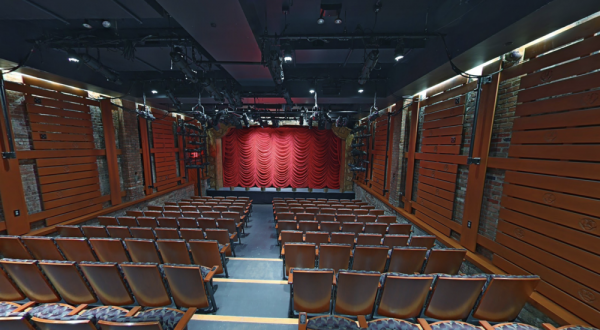 Rows of brown seats face a red-curtained stage in a small West Village theater with wooden walls, brick, and stage lighting.