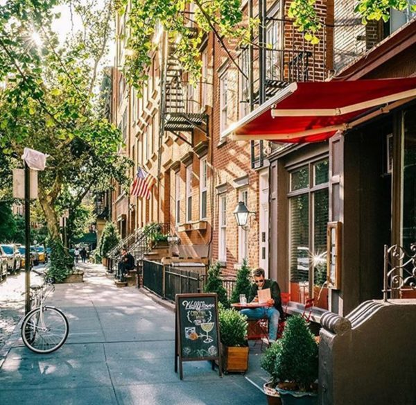 In the West Village, a person works on a laptop at a sidewalk café shaded by trees; an American flag hangs nearby.