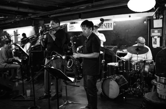 A jazz band plays trombone, saxophone, piano, bass, and drums onstage in a dim West Village club in black and white.