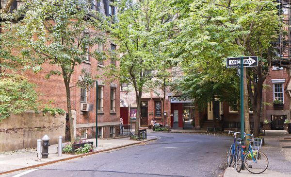 A curved, tree-shaded street in the West Village shows red brick buildings, benches, a one-way sign, and two bicycles.