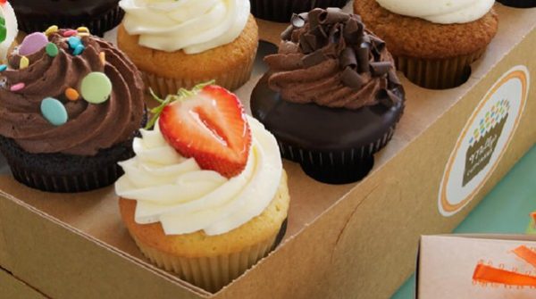 A box of cupcakes with bright frosting, sprinkles, and fruit sits next to a small gift box tied with an orange ribbon.