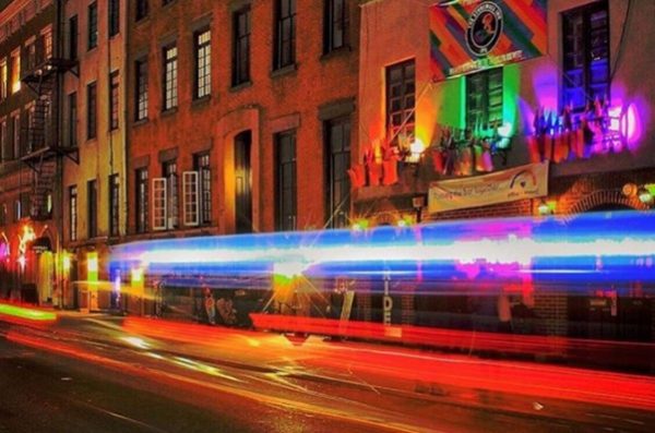 Colorful light trails pass a brick building with rainbow flags and neon lights on a lively West Village street at night.